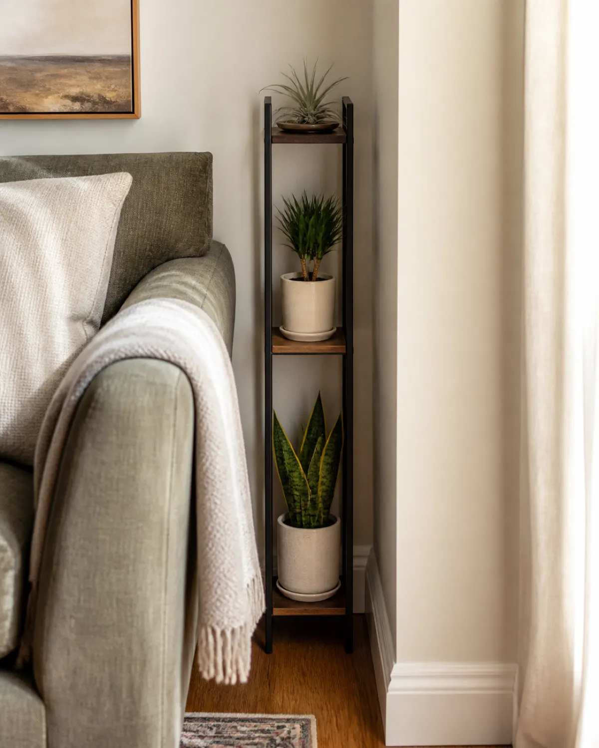 A very narrow tall plant stand about eight inches wide fitting in the gap between a sofa arm and wall holding three small upright plants a compact snake plant a small dracaena and an air plant tight space emphasized warm living room natural side light